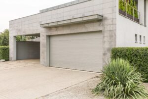 Modern house exterior with a closed light gray garage door, tiled driveway, and green bushes in the foreground.