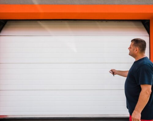 A man stands in front of a closed white garage door with an orange frame, holding a remote control and facing the door.