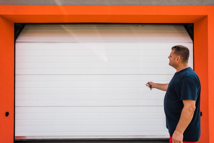 A man stands in front of a closed white garage door with an orange frame, holding a remote control and facing the door.