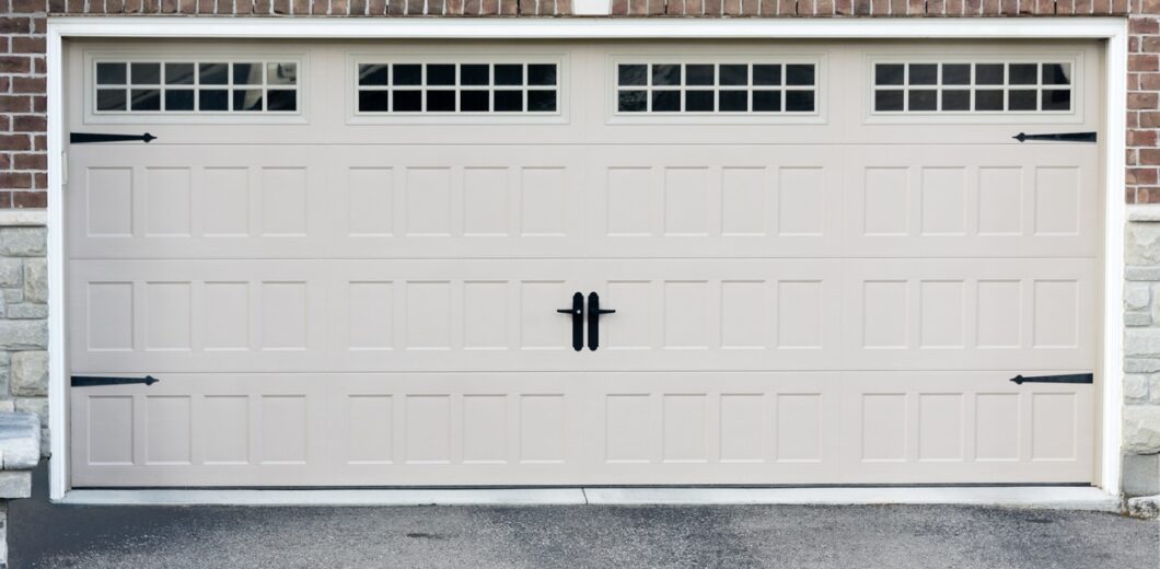 Closed beige garage door with three rectangular windows at the top, black handles in the center, and black decorative hardware, set in a brick and stone exterior wall.