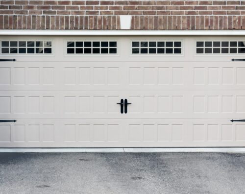 Closed beige garage door with three rectangular windows at the top, black handles in the center, and black decorative hardware, set in a brick and stone exterior wall.