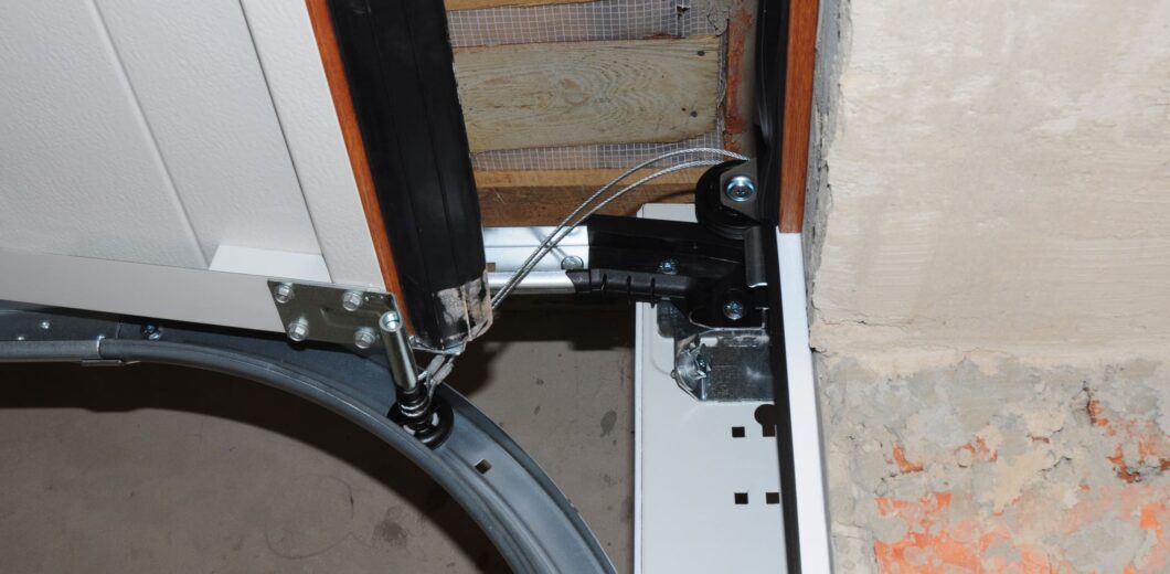 Close-up of a garage door mechanism showing the metal track, cable, hinge, and part of the unfinished wall with exposed bricks.