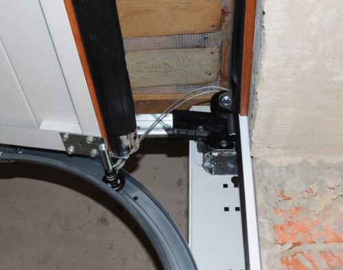 Close-up of a garage door mechanism showing the metal track, cable, hinge, and part of the unfinished wall with exposed bricks.