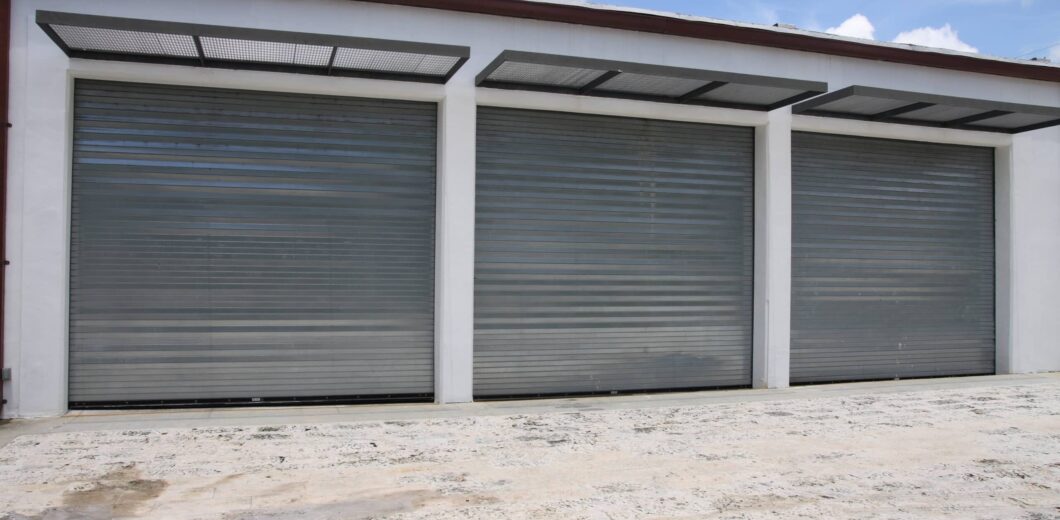 Three closed metallic roller garage doors set in a white building, with a concrete ground and a blue sky with clouds above.