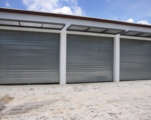 Three closed metallic roller garage doors set in a white building, with a concrete ground and a blue sky with clouds above.