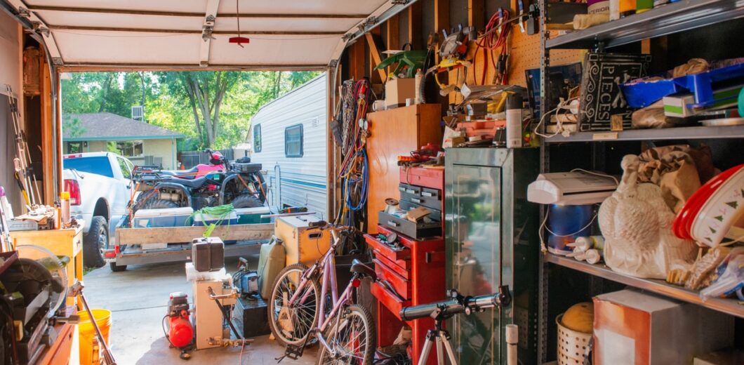 A cluttered garage with tools, bikes, shelves full of various items, and an open door showing vehicles and a trailer outside.