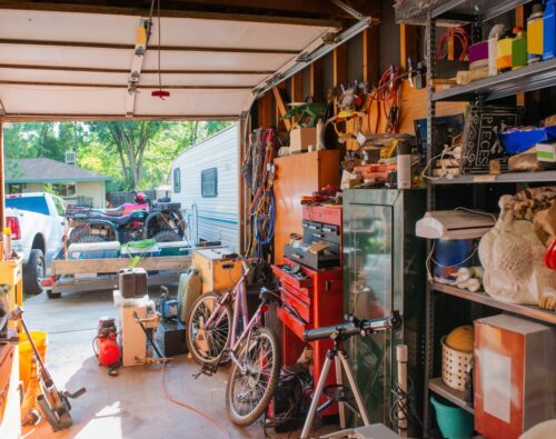 A cluttered garage with tools, bikes, shelves full of various items, and an open door showing vehicles and a trailer outside.