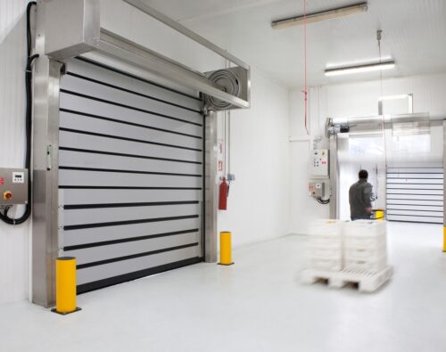 A person pushes a pallet jack with stacked trays through a clean, industrial loading area with large automated doors and white walls.