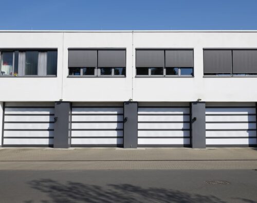 Four modern white garage doors with dark frames are aligned side by side in a white building, each with a window above, under a clear blue sky.