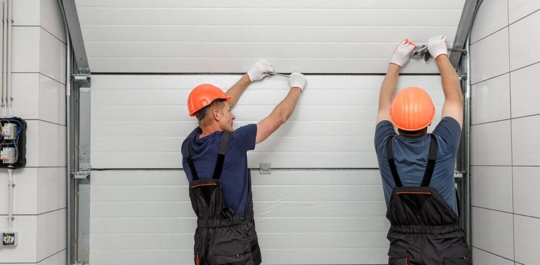 Two workers in safety helmets and overalls install or repair a sectional garage door from the inside of a building.