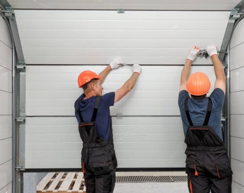 Two workers in safety helmets and overalls install or repair a sectional garage door from the inside of a building.