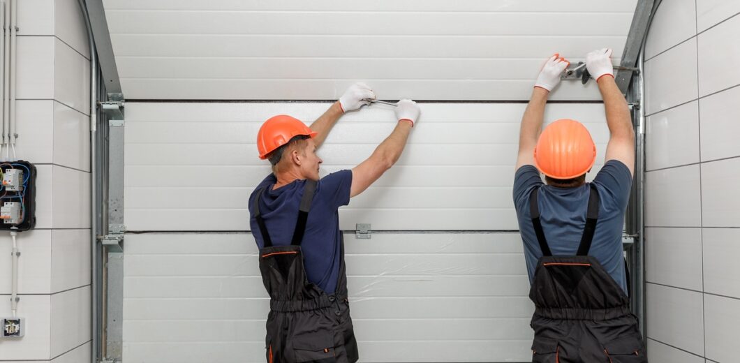 Two workers in orange hard hats and overalls installing a white sectional garage door from inside a garage.