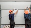 Two workers in orange hard hats and overalls installing a white sectional garage door from inside a garage.