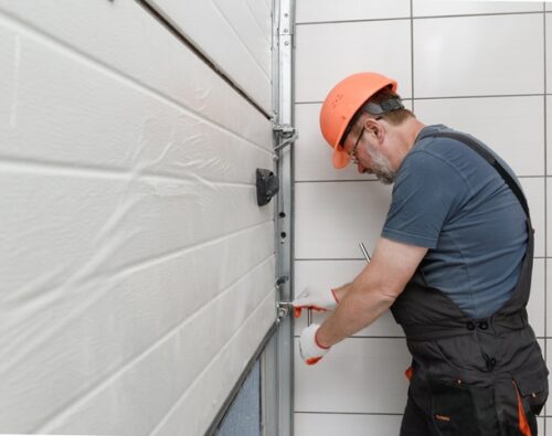 A man in work overalls and an orange hard hat repairs or installs a garage door, using a screwdriver on the metal frame next to a tiled wall.