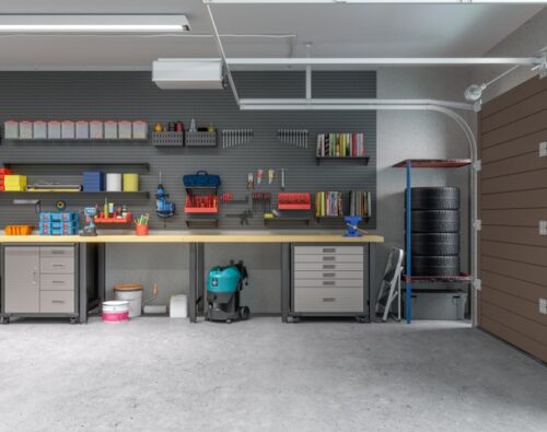 A clean, organized garage with workbenches, storage cabinets, tools on wall panels, a stack of tires, a vacuum, and the garage door partially open.
