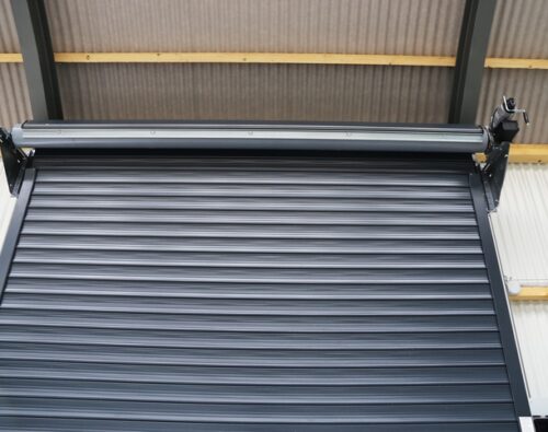 A metal roller shutter door is partially open inside an industrial building with a corrugated roof and metal frame.