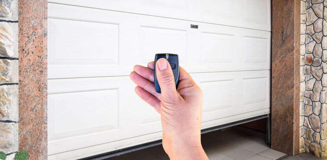 A hand holding a remote control in front of a partially opened white garage door with a stone and tile exterior.