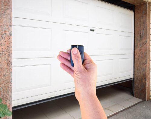 A hand holding a remote control in front of a partially opened white garage door with a stone and tile exterior.