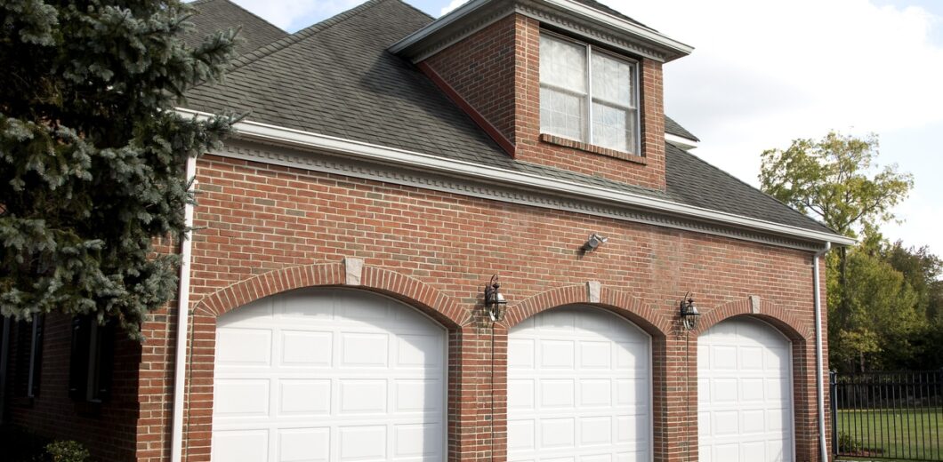 A red brick house with three white garage doors, exterior wall lamps, a dormer window above, and a black paved driveway in front.