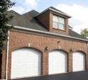 A red brick house with three white garage doors, exterior wall lamps, a dormer window above, and a black paved driveway in front.