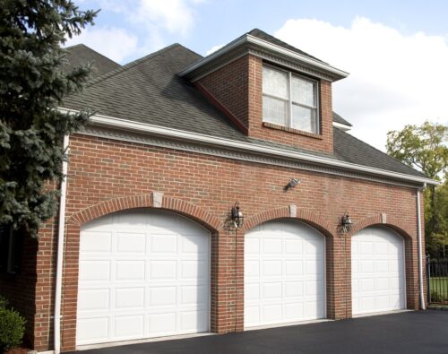 A red brick house with three white garage doors, exterior wall lamps, a dormer window above, and a black paved driveway in front.