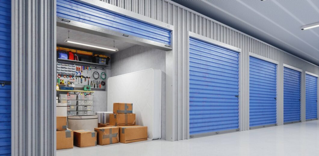 A row of storage units with blue doors; one unit is open, showing boxes, a mattress, and various tools and supplies organized on shelves.