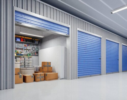 A row of storage units with blue doors; one unit is open, showing boxes, a mattress, and various tools and supplies organized on shelves.