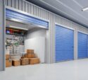 A row of storage units with blue doors; one unit is open, revealing cardboard boxes, a mattress, and organized tools hanging on the back wall.