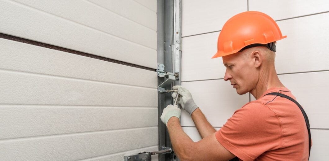 A worker in an orange hard hat adjusts a metal bracket on a garage door using a wrench.