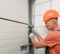 A worker in an orange hard hat adjusts a metal bracket on a garage door using a wrench.
