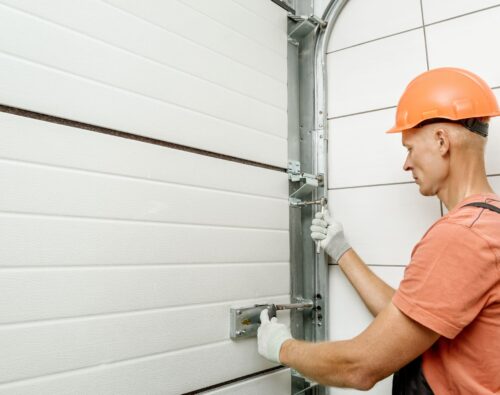 A worker in a hard hat and gloves is installing or repairing a sectional garage door, adjusting the metal tracks and hinges with a tool.