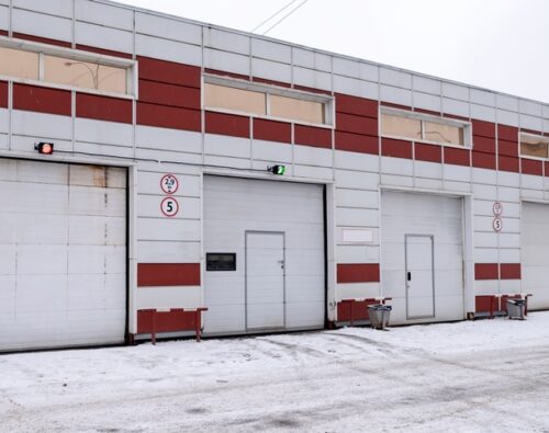 A row of industrial garage doors in a building with red and white panels, one door showing a green light, others red, with snow on the ground.