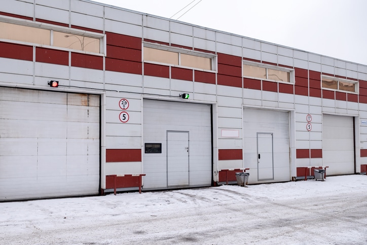 A row of industrial garage doors in a building with red and white panels, one door showing a green light, others red, with snow on the ground.