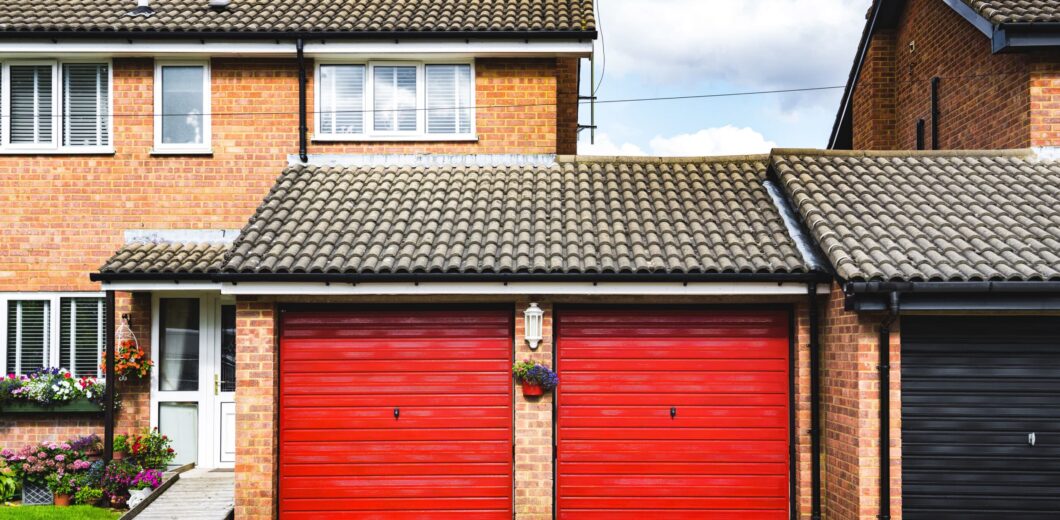 Two red garage doors attached to a brick house with a tiled roof, a gravel driveway, and a small hanging flower basket between the doors.
