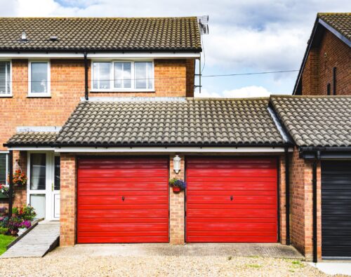 Two red garage doors attached to a brick house with a tiled roof, a gravel driveway, and a small hanging flower basket between the doors.