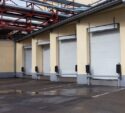 Four closed white loading dock doors on the side of an industrial building with pipes overhead and damp pavement in front.