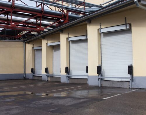 Four closed white loading dock doors on the side of an industrial building with pipes overhead and damp pavement in front.