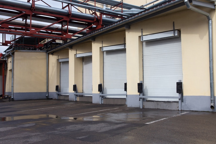 Four closed white loading dock doors on the side of an industrial building with pipes overhead and damp pavement in front.