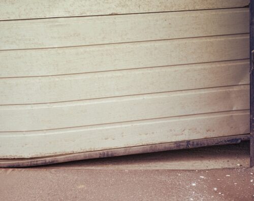 A garage door is dented and bent at the bottom, with visible dirt on the ground in front of it.