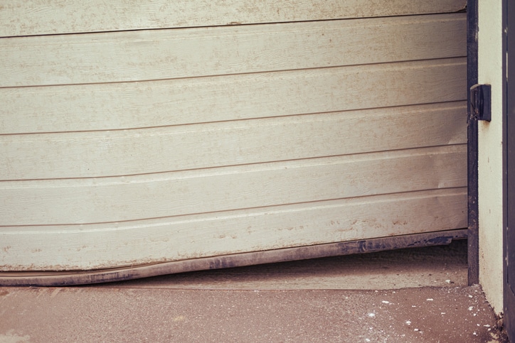 A garage door is dented and bent at the bottom, with visible dirt on the ground in front of it.