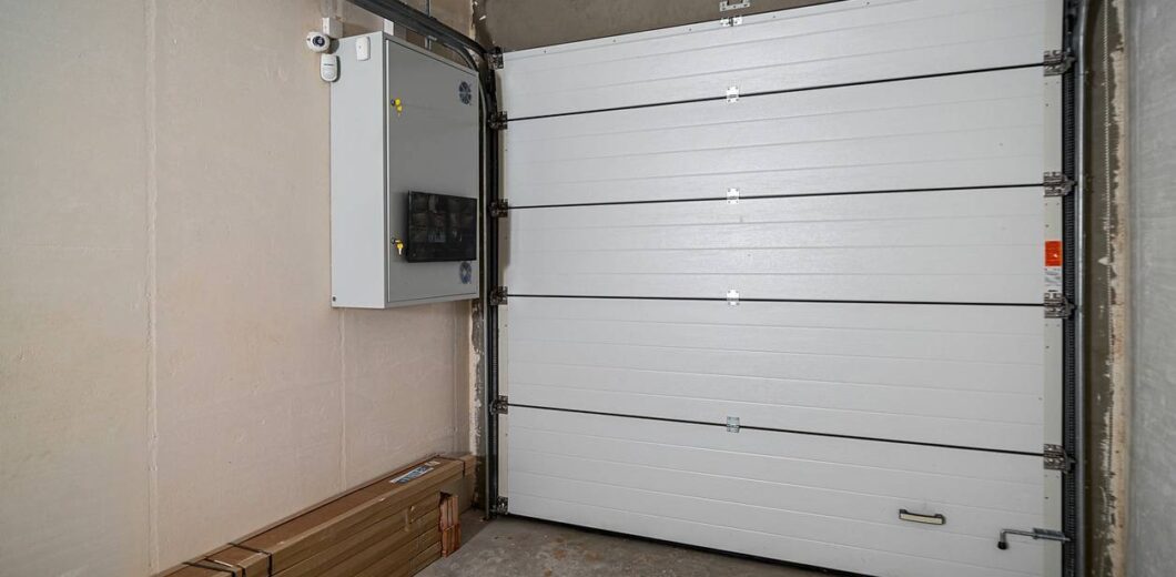 Closed white sectional garage door in a bare concrete garage with an electrical panel and some long, boxed items on the floor.