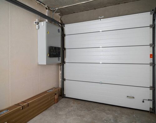 Closed white sectional garage door in a bare concrete garage with an electrical panel and some long, boxed items on the floor.