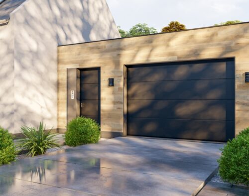 Modern detached garage with light wood siding, a dark single garage door, side entry door, two wall lights, and landscaped bushes on a concrete driveway.