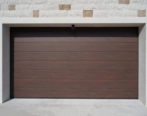 Closed brown garage door with horizontal panels set in a light-colored brick wall with a small exterior light above the door.