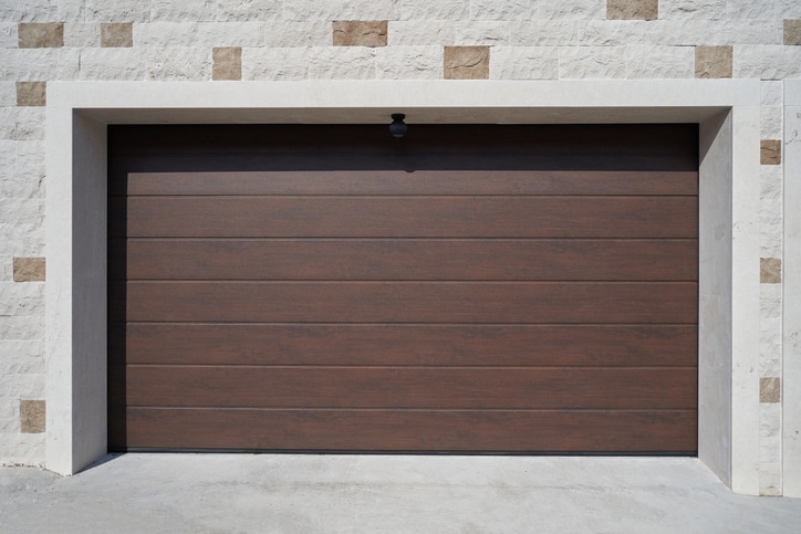 Closed brown garage door with horizontal panels set in a light-colored brick wall with a small exterior light above the door.