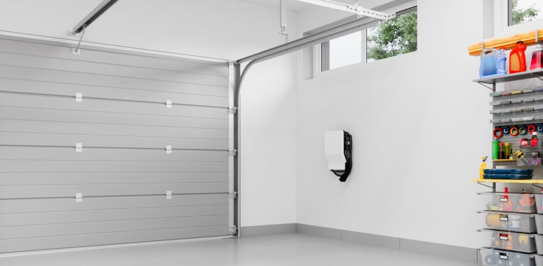 Clean, empty garage with a closed gray door, white walls, high windows, a wall-mounted device, and organized shelves with tools and supplies on the right.