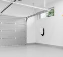Clean, empty garage with a closed gray door, white walls, high windows, a wall-mounted device, and organized shelves with tools and supplies on the right.