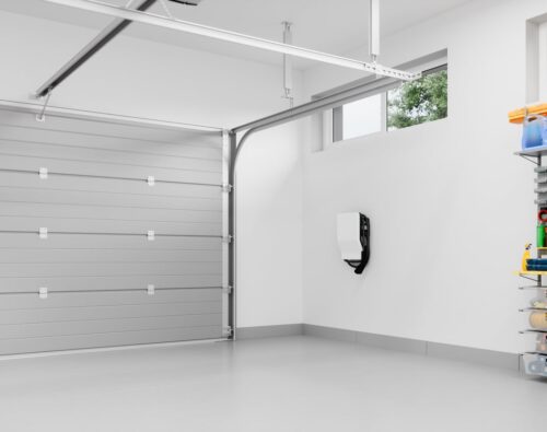 Clean, empty garage with a closed gray door, white walls, high windows, a wall-mounted device, and organized shelves with tools and supplies on the right.