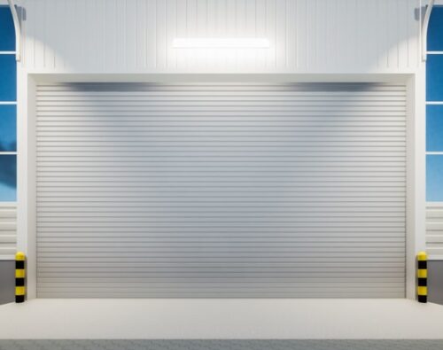 Closed white industrial garage door with protective yellow-and-black striped bollards on either side, illuminated by a wall-mounted light and surrounded by large windows.