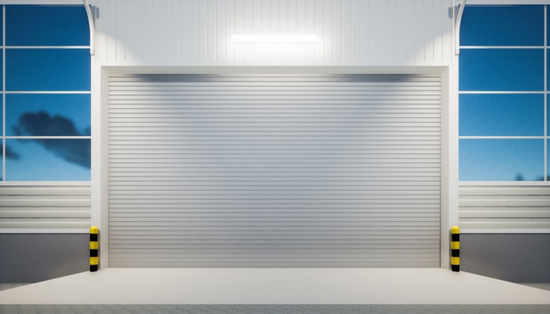Closed white industrial garage door with protective yellow-and-black striped bollards on either side, illuminated by a wall-mounted light and surrounded by large windows.